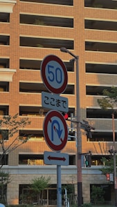 A street scene featuring traffic signs in front of a multi-story building with a brick facade. The signs indicate speed limit and a no U-turn restriction. Trees and traffic lights are also present, with sunlight casting shadows on the building.
