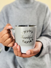A person wearing a gray sweater is holding a light gray mug with the word 'Thankful' written on it, decorated with a leaf motif. The mug is being held with both hands, implying a sense of warmth and comfort.