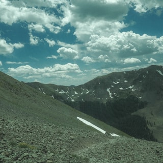 A rocky mountain landscape with partially snow-covered peaks and a bright blue sky filled with fluffy white clouds.