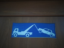 A pictogram on a blue background depicts a tow truck pulling a car. The design is simple with the vehicles shown in white outlines against a vibrant blue rectangle. Both vehicles are oriented in a profile view, clearly illustrating the towing action.