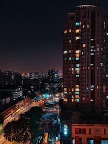 A nighttime cityscape featuring a tall building illuminated by various colored lights, with a busy street below. The street is lined with trees and smaller buildings, and there are visible signs of ongoing construction. The distant skyline is visible against the night sky.