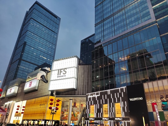 A cityscape featuring modern high-rise buildings with large glass windows. Prominent signage for luxury brands like Prada, Valentino, and Ferragamo is visible, alongside a significant display of a giant panda climbing one of the buildings. Evening light reflects off the buildings, and decorative red lanterns suggest a festive atmosphere.