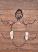 A person is positioned face-forward through a wooden board with holes for the head, hands, and feet. Chains connect the hands and feet, resembling stocks or a restraint setup. The wood has a natural, polished texture with a warm brown color.