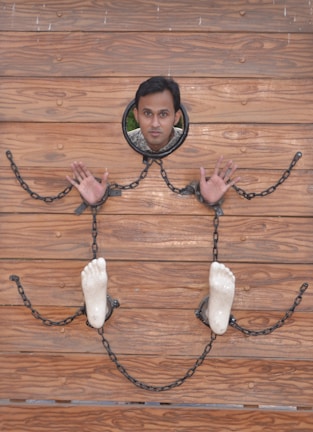 A person is positioned face-forward through a wooden board with holes for the head, hands, and feet. Chains connect the hands and feet, resembling stocks or a restraint setup. The wood has a natural, polished texture with a warm brown color.