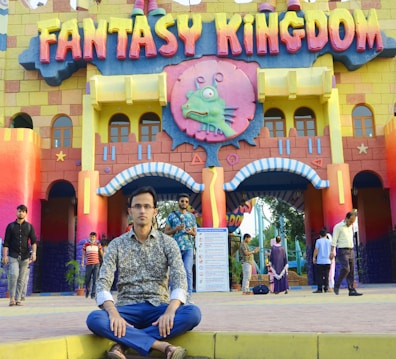 A young man sits cross-legged in front of a colorful, castle-like entrance adorned with the words 'Fantasy Kingdom'. The building features vibrant yellow, red, and blue hues, with playful elements and a cartoonish monster icon above the entrance. Several other people are walking around the area, giving a lively and bustling atmosphere.