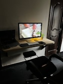 A modern workspace with a desktop computer on a wooden wall-mounted desk. The screen displays a close-up of hands working with a piece of material. A small potted plant is beside the monitor, alongside a keyboard and mouse. A laptop is partially visible to the left. An office chair is positioned in front of the desk, and a wooden door is in the background.