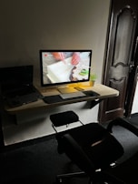 A modern workspace with a desktop computer on a wooden wall-mounted desk. The screen displays a close-up of hands working with a piece of material. A small potted plant is beside the monitor, alongside a keyboard and mouse. A laptop is partially visible to the left. An office chair is positioned in front of the desk, and a wooden door is in the background.