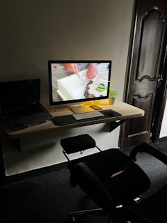A modern workspace with a desktop computer on a wooden wall-mounted desk. The screen displays a close-up of hands working with a piece of material. A small potted plant is beside the monitor, alongside a keyboard and mouse. A laptop is partially visible to the left. An office chair is positioned in front of the desk, and a wooden door is in the background.