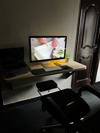 A modern workspace with a desktop computer on a wooden wall-mounted desk. The screen displays a close-up of hands working with a piece of material. A small potted plant is beside the monitor, alongside a keyboard and mouse. A laptop is partially visible to the left. An office chair is positioned in front of the desk, and a wooden door is in the background.