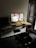 A modern workspace with a desktop computer on a wooden wall-mounted desk. The screen displays a close-up of hands working with a piece of material. A small potted plant is beside the monitor, alongside a keyboard and mouse. A laptop is partially visible to the left. An office chair is positioned in front of the desk, and a wooden door is in the background.