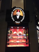 A shop display featuring an ornate sign with a portrait of a man in historical attire and text above and below the image. Below the sign, a brightly lit showcase displays a variety of desserts and confections, with pyramids of sweets and packaged items arranged on shelves. The background and accents are primarily red, enhancing the opulent presentation.