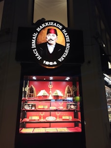 A shop display featuring an ornate sign with a portrait of a man in historical attire and text above and below the image. Below the sign, a brightly lit showcase displays a variety of desserts and confections, with pyramids of sweets and packaged items arranged on shelves. The background and accents are primarily red, enhancing the opulent presentation.