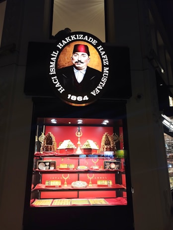 A shop display featuring an ornate sign with a portrait of a man in historical attire and text above and below the image. Below the sign, a brightly lit showcase displays a variety of desserts and confections, with pyramids of sweets and packaged items arranged on shelves. The background and accents are primarily red, enhancing the opulent presentation.