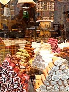 A colorful display of various types of Turkish delight stacked in pyramids behind a glass window. The confections are arranged in different flavors, including ones with pistachios, almonds, and assorted coatings. The setting appears to be a vibrant marketplace or a specialty shop with shelves filled with boxed products and decorative lighting in the background.
