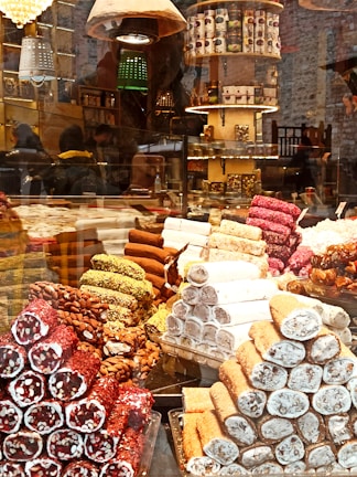 A colorful display of various types of Turkish delight stacked in pyramids behind a glass window. The confections are arranged in different flavors, including ones with pistachios, almonds, and assorted coatings. The setting appears to be a vibrant marketplace or a specialty shop with shelves filled with boxed products and decorative lighting in the background.