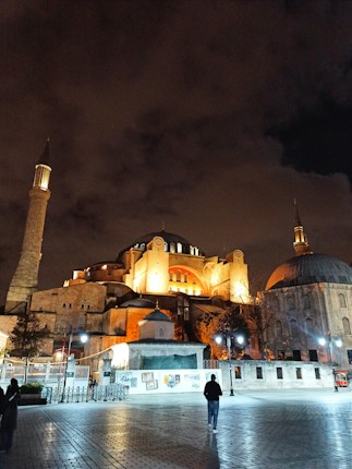 A historic, illuminated mosque with architectural features, including a prominent dome and minarets, set against a dramatic evening sky. The scene shows a person walking in a large, open courtyard paved with stones.