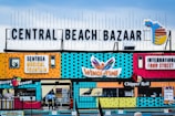 A colorful and vibrant storefront displays bold signage with a variety of attractions and food outlets. The large text reading 'Central Beach Bazaar' is prominently featured at the top, accompanied by signage for 'Sentosa Musical Fountain,' 'Wings of Time,' and 'International Food Street.' The building exterior is decorated with playful patterns and bright colors, creating an inviting atmosphere.