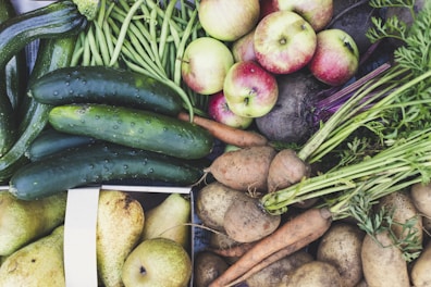 An abundant assortment of fresh vegetables and fruits including cucumbers, green beans, apples, carrots, potatoes, and pears. The produce is vibrant and appears freshly harvested, with earthy tones and organic textures.