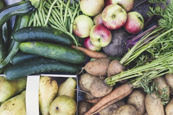 An abundant assortment of fresh vegetables and fruits including cucumbers, green beans, apples, carrots, potatoes, and pears. The produce is vibrant and appears freshly harvested, with earthy tones and organic textures.