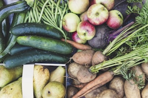 An abundant assortment of fresh vegetables and fruits including cucumbers, green beans, apples, carrots, potatoes, and pears. The produce is vibrant and appears freshly harvested, with earthy tones and organic textures.
