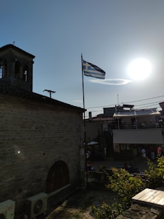 A Greek flag flutters atop a pole beside an old stone building with a bell tower, silhouetted against a bright sun. In the background, a modern structure is visible, and people are walking on the street below. The scene captures a blend of historical architecture and contemporary life.