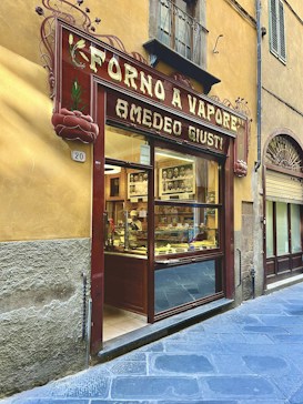 The image depicts a traditional bakery storefront with a vintage wooden sign displaying the name 'Forno A Vapore Amedeo Giusti.' The shop is located on a quaint street with stone pavement and is set in a building with rustic yellow plaster walls. Inside the bakery, shelves are filled with various baked goods, and the interior is softly illuminated, creating a warm and inviting atmosphere.