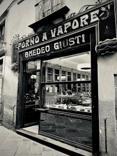 A vintage storefront with a sign reading 'Forno a Vapore Amedeo Giusti' above the entrance. The exterior features decorative elements and large glass windows displaying a variety of baked goods. The building is situated on a stone-paved street and has an old-fashioned charm.