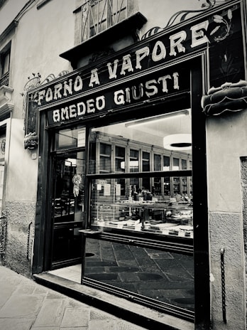 A vintage storefront with a sign reading 'Forno a Vapore Amedeo Giusti' above the entrance. The exterior features decorative elements and large glass windows displaying a variety of baked goods. The building is situated on a stone-paved street and has an old-fashioned charm.