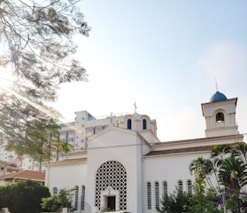 A white church building with classic architectural features, such as arched windows and a decorative facade. The structure is surrounded by lush greenery and is set against a bright, clear sky. Sunlight filters through the branches of a nearby tree, casting dappled light across the scene.