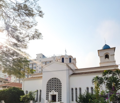A white church building with classic architectural features, such as arched windows and a decorative facade. The structure is surrounded by lush greenery and is set against a bright, clear sky. Sunlight filters through the branches of a nearby tree, casting dappled light across the scene.