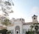 A white church building with classic architectural features, such as arched windows and a decorative facade. The structure is surrounded by lush greenery and is set against a bright, clear sky. Sunlight filters through the branches of a nearby tree, casting dappled light across the scene.
