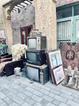 An outdoor vintage scene featuring a stack of old televisions, framed pictures, and various antique items. The televisions are stacked on top of each other, surrounded by a collection of eclectic objects including a samovar, a wooden spool, and paintings. The backdrop consists of an old textured wall with a partially visible archway. The area seems to exude a sense of nostalgia.