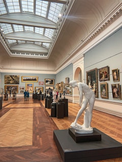 An art gallery with a high, arched ceiling and skylights allowing natural light to illuminate the space. Numerous framed paintings hang on the walls, showcasing various styles. Sculptures are positioned on pedestals throughout the gallery. Visitors are observing and walking through the space.