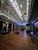 A grand hall with high ceilings featuring ornate architectural details. There is a skeletal dinosaur display hanging from the ceiling in the center of the room and another skeletal display on the left. People are walking on the polished wooden floor, and glass cases line the walls, possibly displaying various artifacts.