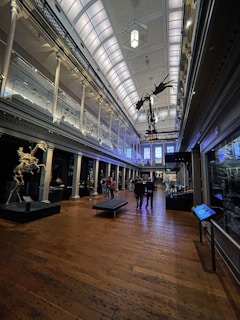 A grand hall with high ceilings featuring ornate architectural details. There is a skeletal dinosaur display hanging from the ceiling in the center of the room and another skeletal display on the left. People are walking on the polished wooden floor, and glass cases line the walls, possibly displaying various artifacts.
