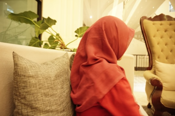 A person wearing a red hijab sits on a light-colored sofa with a textured gray pillow. A large green plant with broad leaves is nearby, and a tufted armchair is visible in the background. The setting appears comfortable and indoors.