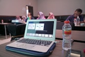 A laptop displaying a presentation is on a wooden table alongside a water bottle. In the background, several people are sitting in a conference room, some wearing headscarves. A projector is also visible, projecting content onto an unseen screen.