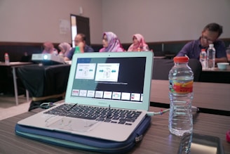 A laptop displaying a presentation is on a wooden table alongside a water bottle. In the background, several people are sitting in a conference room, some wearing headscarves. A projector is also visible, projecting content onto an unseen screen.