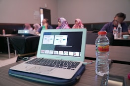 A laptop displaying a presentation is on a wooden table alongside a water bottle. In the background, several people are sitting in a conference room, some wearing headscarves. A projector is also visible, projecting content onto an unseen screen.