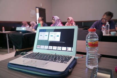 A laptop displaying a presentation is on a wooden table alongside a water bottle. In the background, several people are sitting in a conference room, some wearing headscarves. A projector is also visible, projecting content onto an unseen screen.
