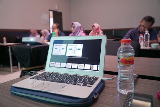 A laptop displaying a presentation is on a wooden table alongside a water bottle. In the background, several people are sitting in a conference room, some wearing headscarves. A projector is also visible, projecting content onto an unseen screen.