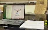 A modern workspace featuring a laptop displaying an email application. The screen shows a message notification from LinkedIn. Adjacent to the laptop, a stack of papers with highlighted text and a pair of glasses are visible. A yellow highlighter rests beside the papers. The background includes a blurred office chair.
