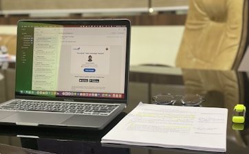 A modern workspace featuring a laptop displaying an email application. The screen shows a message notification from LinkedIn. Adjacent to the laptop, a stack of papers with highlighted text and a pair of glasses are visible. A yellow highlighter rests beside the papers. The background includes a blurred office chair.