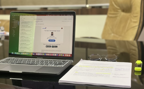 A modern workspace featuring a laptop displaying an email application. The screen shows a message notification from LinkedIn. Adjacent to the laptop, a stack of papers with highlighted text and a pair of glasses are visible. A yellow highlighter rests beside the papers. The background includes a blurred office chair.