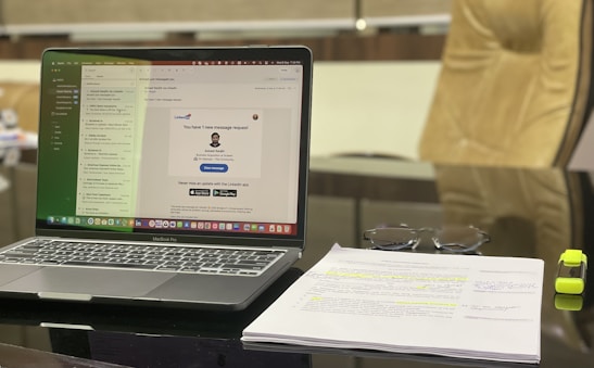 A modern workspace featuring a laptop displaying an email application. The screen shows a message notification from LinkedIn. Adjacent to the laptop, a stack of papers with highlighted text and a pair of glasses are visible. A yellow highlighter rests beside the papers. The background includes a blurred office chair.