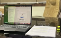 A modern workspace featuring a laptop displaying an email application. The screen shows a message notification from LinkedIn. Adjacent to the laptop, a stack of papers with highlighted text and a pair of glasses are visible. A yellow highlighter rests beside the papers. The background includes a blurred office chair.