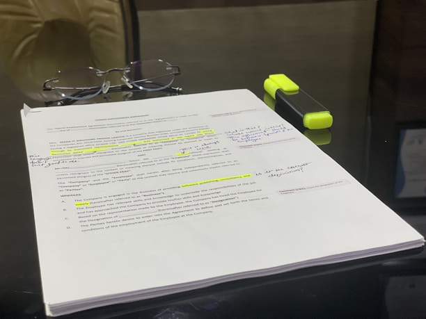 A pair of eyeglasses is placed on top of a stack of printed documents, which have several handwritten notes and highlighted sections. A yellow highlighter pen is next to the papers, lying on a reflective black surface.