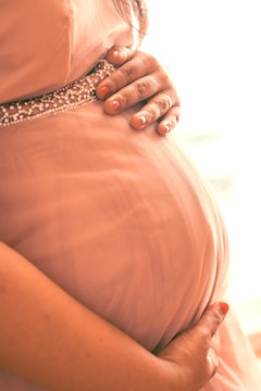 A close-up of a pregnant woman's belly, with hands gently resting on it. The skin is covered by a soft pink dress adorned with a beaded band just below the chest.