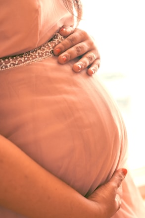 A close-up of a pregnant woman's belly, with hands gently resting on it. The skin is covered by a soft pink dress adorned with a beaded band just below the chest.