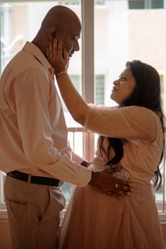 A couple stands in a warmly lit room near a window. The woman, wearing a peach-colored dress, lovingly cups the face of the man, who is dressed in a light-colored shirt and trousers. Their expressions reflect a moment of tenderness and intimacy, with the man's hand placed gently on the woman's pregnant belly.